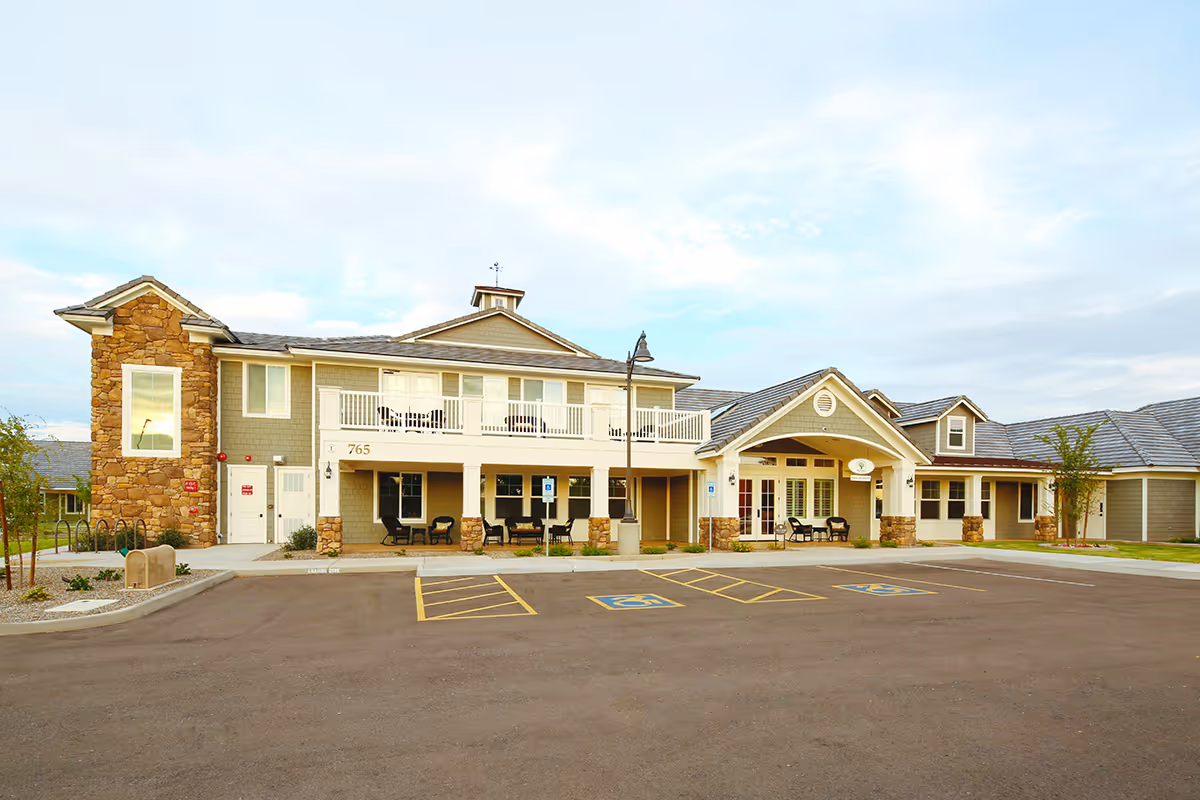 Exterior view of a two-story assisted living and memory care facility building with stone and beige siding, a covered entrance with seating, a balcony, and a parking lot with handicap spaces in front.