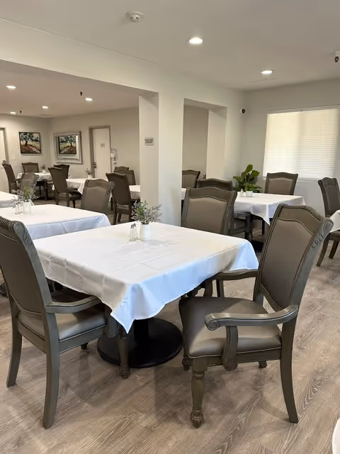 A dining area with multiple tables covered in white tablecloths, each surrounded by cushioned wooden chairs. The room has light-colored walls, wood flooring, and framed artwork on the walls. There are small plants and salt and pepper shakers on the tables, and a large window with blinds letting in natural light.