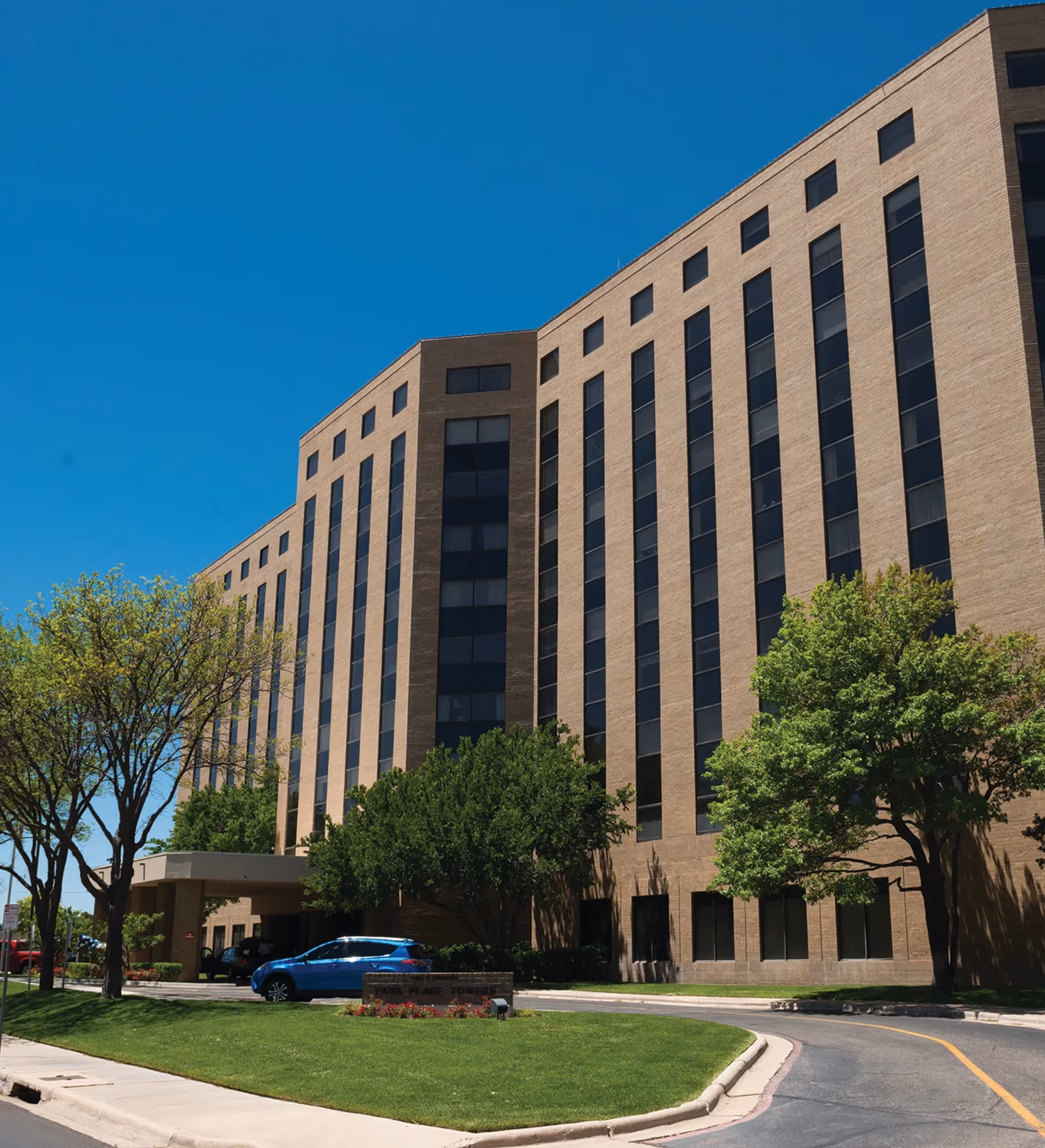 Exterior view of a multi-story brick building with many windows, surrounded by green trees and a well-maintained lawn under a clear blue sky. A blue car is parked near the entrance, and a sign in front reads 'Park Place Towers'.