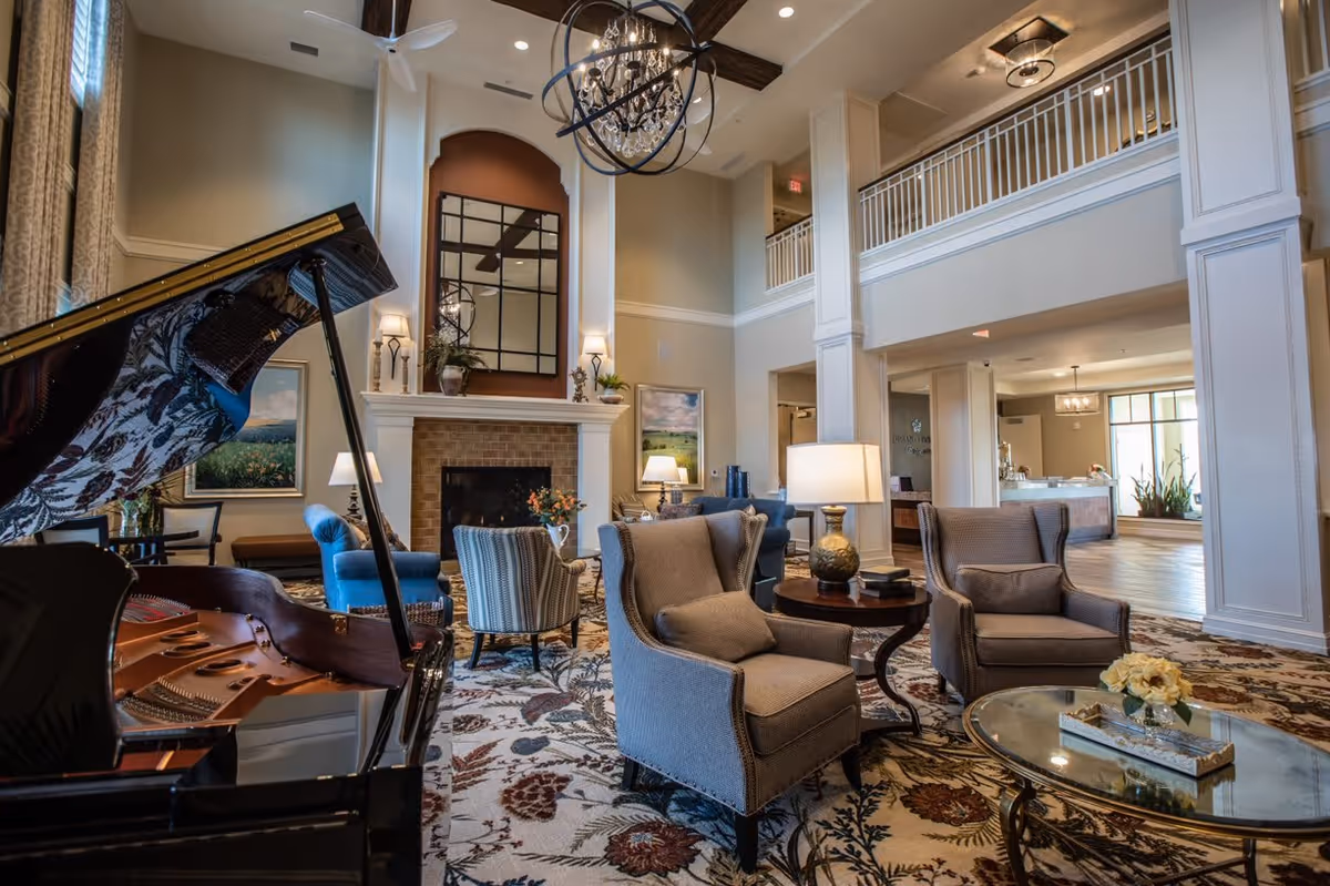 A spacious and elegant living room area with high ceilings, a grand piano, comfortable armchairs, a fireplace, and decorative lighting. The room features a patterned carpet, large windows with curtains, and a balcony overlooking the space.