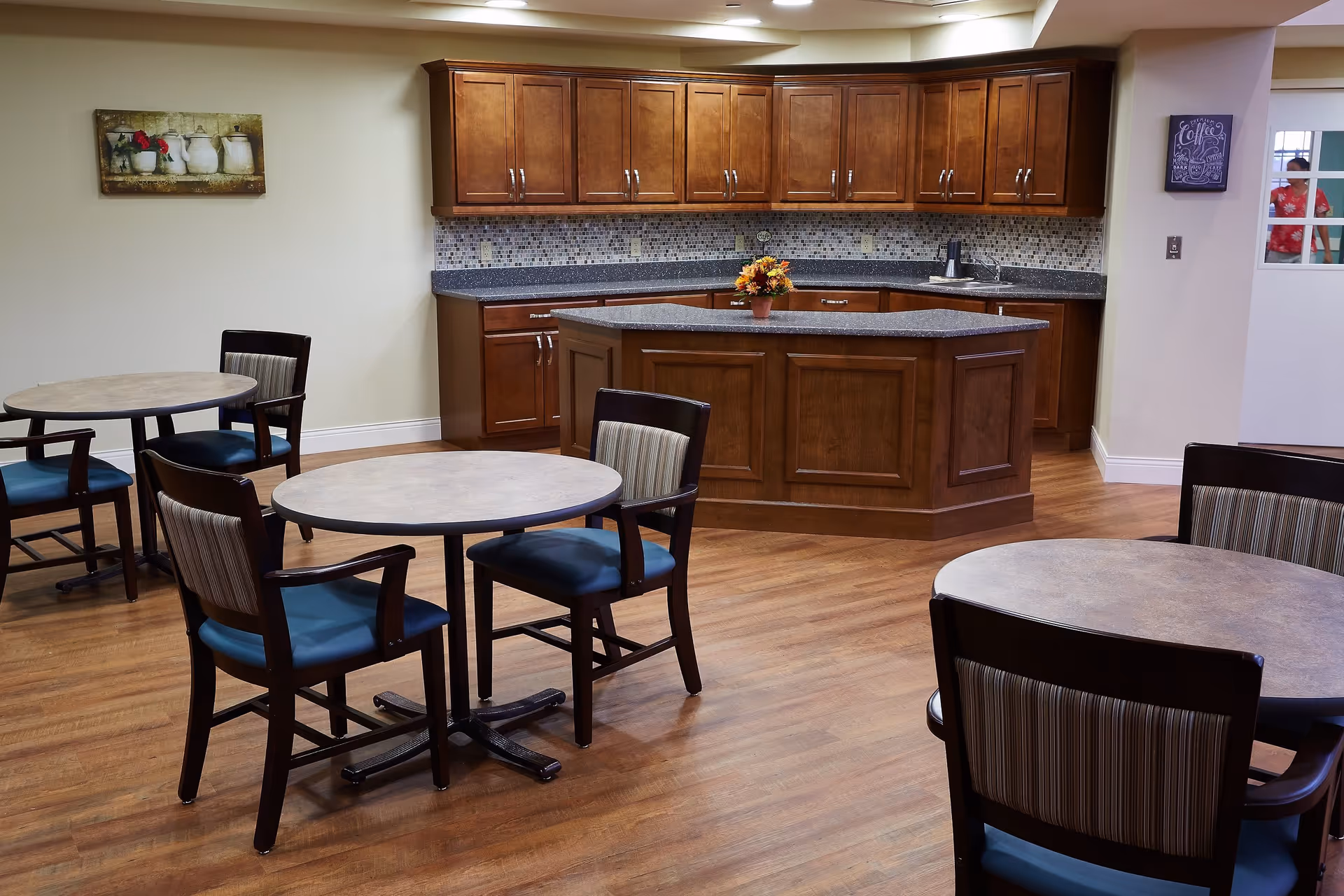 A dining area with round tables and chairs with blue cushions and striped backs, wooden flooring, and a kitchen area with wooden cabinets and a granite countertop island. A small flower arrangement is on the island, and there is a painting on the wall and a coffee-themed sign near a door.