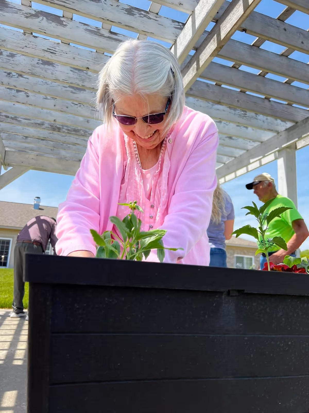 An elderly woman wearing sunglasses and a pink cardigan is smiling while tending to plants in a raised garden bed under a wooden pergola. In the background, two other people are also engaged in gardening activities outside near a building.