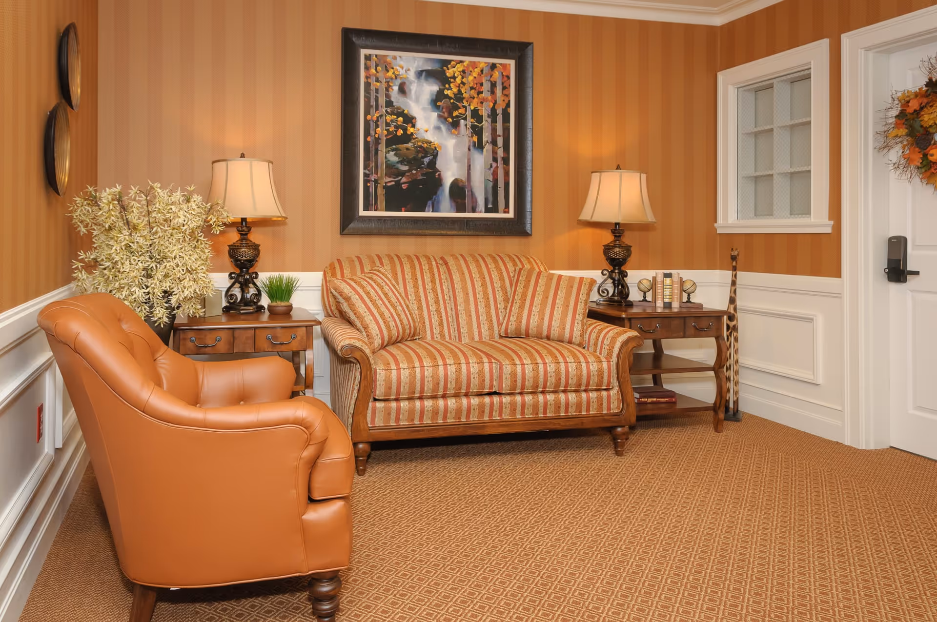 A cozy sitting area with a striped loveseat and a brown leather armchair. There are two wooden side tables with decorative lamps, a small plant, and books. The walls have orange and beige vertical stripes, and a framed painting of a waterfall with autumn trees hangs above the loveseat. The carpet is patterned, and a door with a fall-themed wreath is visible on the right.