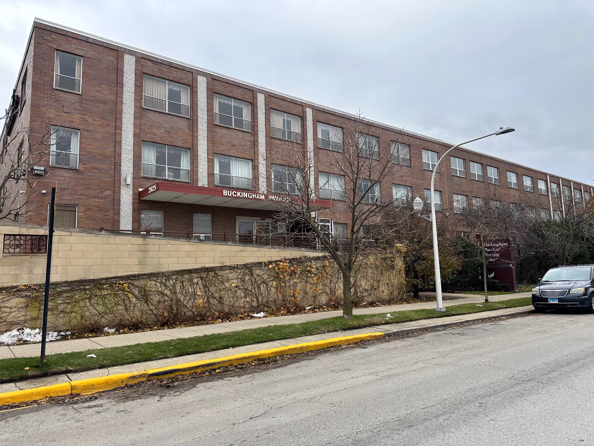 Exterior view of Buckingham Pavilion Nursing and Rehabilitation building, a three-story brick structure with multiple windows. There is a maroon awning above the entrance with the name partially visible. Leafless trees and a sidewalk line the street in front, with a parked car on the right side.