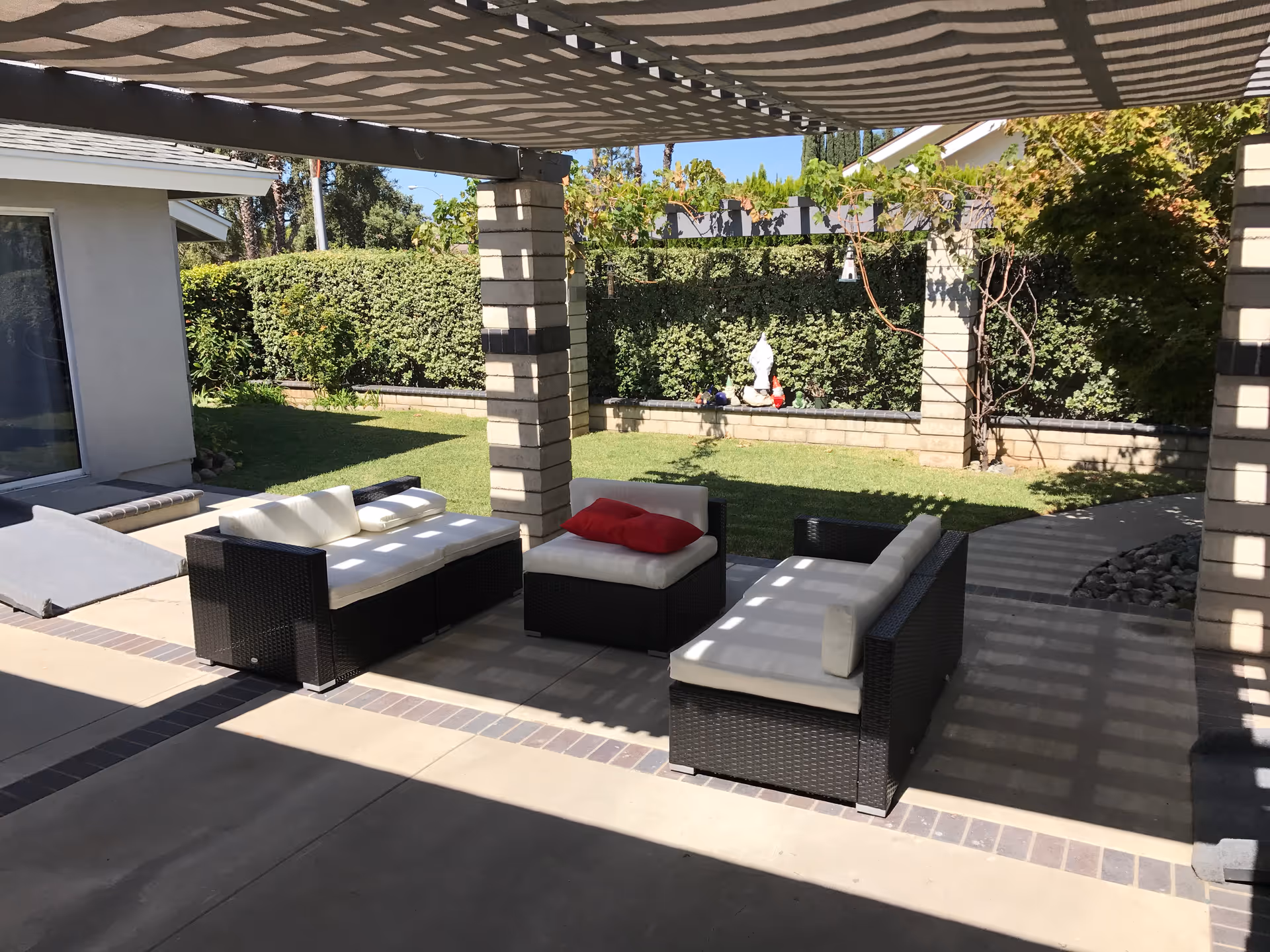 Outdoor patio area with wicker furniture including two cushioned sofas and a cushioned ottoman with a red pillow, shaded by a pergola. The patio overlooks a green lawn bordered by a hedge and a small statue in the background.