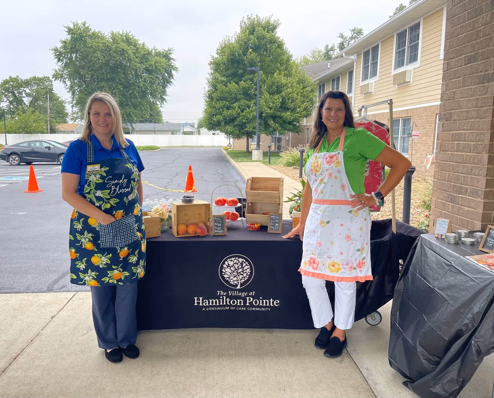 Two women wearing aprons stand outside next to a table covered with a black tablecloth that has The Village at Hamilton Pointe logo and name. The table displays fresh fruits and small wooden crates. Behind them is a parking lot with orange cones and trees.