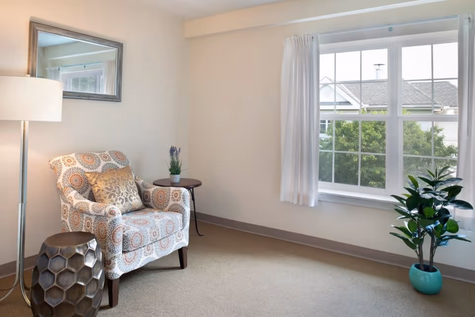A cozy corner of a room featuring a patterned armchair with a decorative pillow, a small round side table with a potted plant, a tall floor lamp, a large window with white curtains, and a green potted plant in a turquoise pot on the floor.