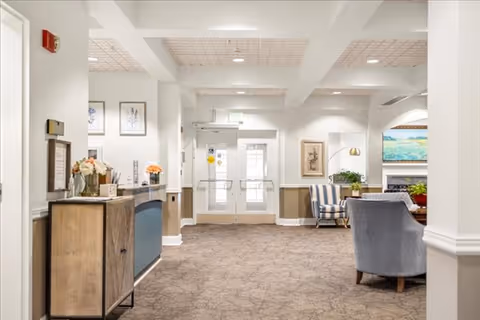 Interior view of a senior living facility lobby or common area with a carpeted floor, white walls, and ceiling with recessed lighting. There is a wooden cabinet with flowers on the left, a reception desk, and seating area with upholstered chairs and framed artwork on the walls. Double glass doors are visible at the far end.