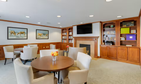Cozy common room with round tables and upholstered chairs, built-in wood bookshelves, a fireplace, and a wall-mounted TV.