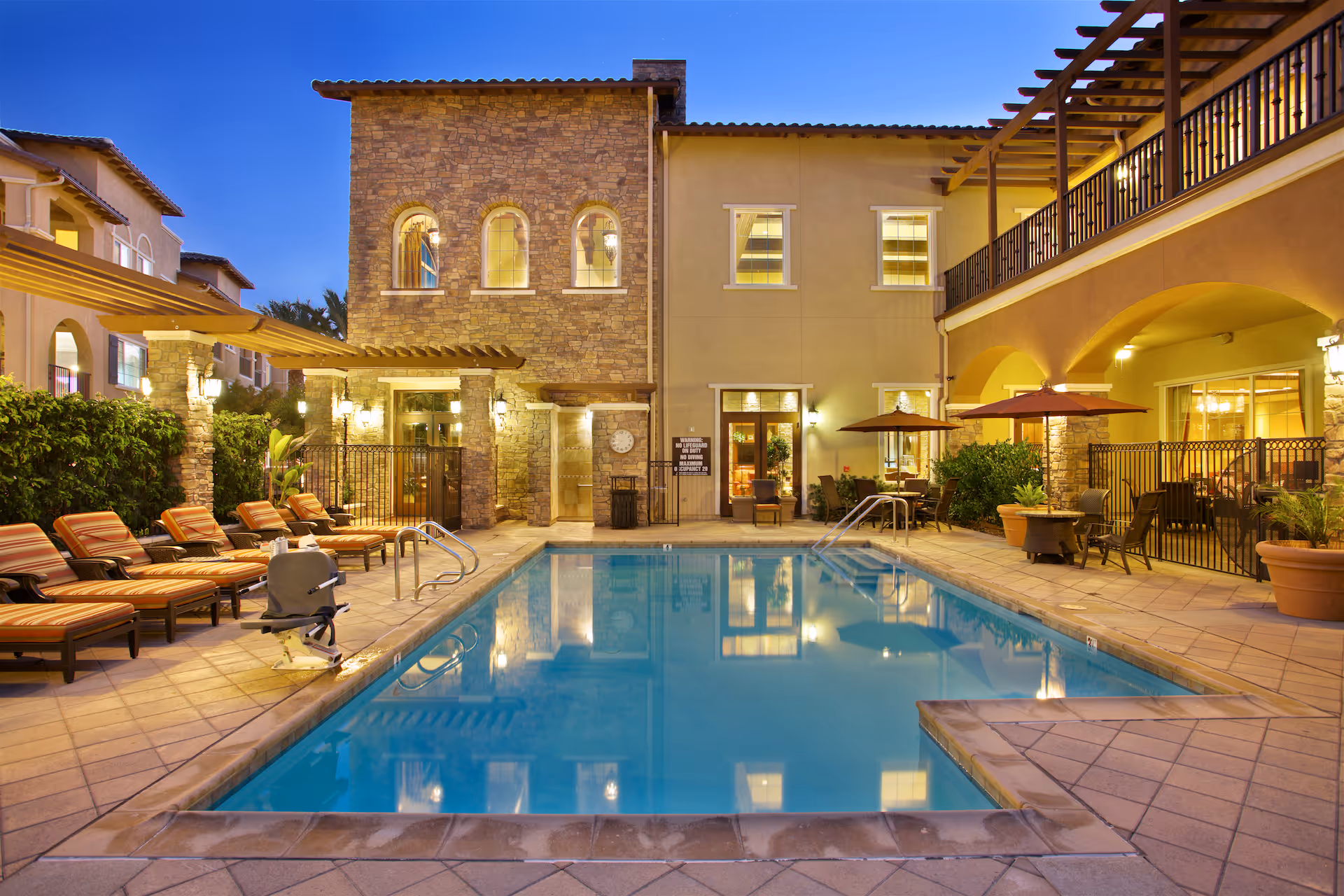 Outdoor courtyard with a swimming pool, lounge chairs, umbrellas, and a multi-story building at dusk.