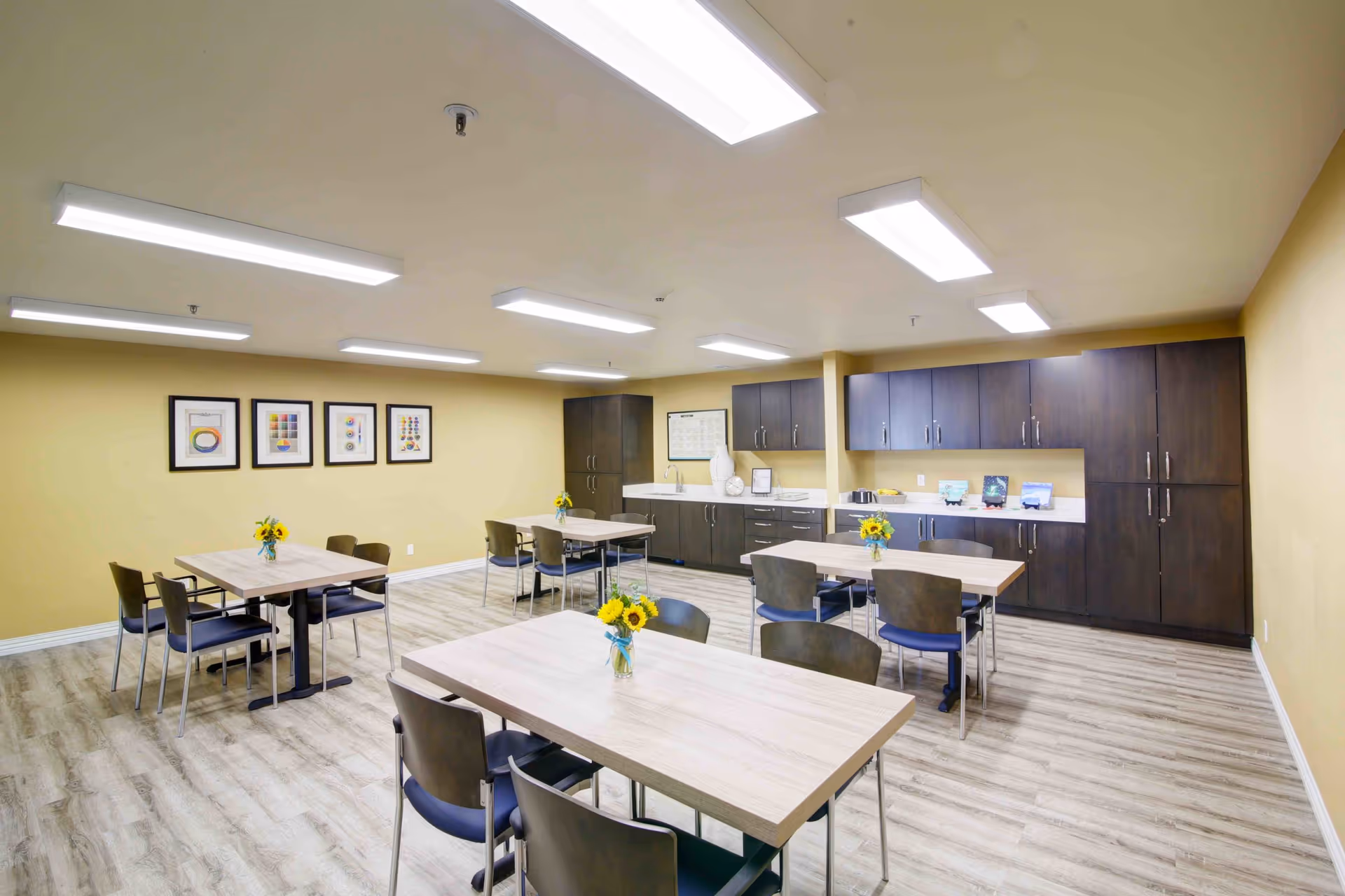 A bright, clean dining area in a senior living facility with light wood tables, dark chairs, and small vases of sunflowers on each table. The room has light yellow walls, wood flooring, and dark wood cabinets along one wall with a countertop and sink. Four framed colorful abstract art pieces hang on one wall.