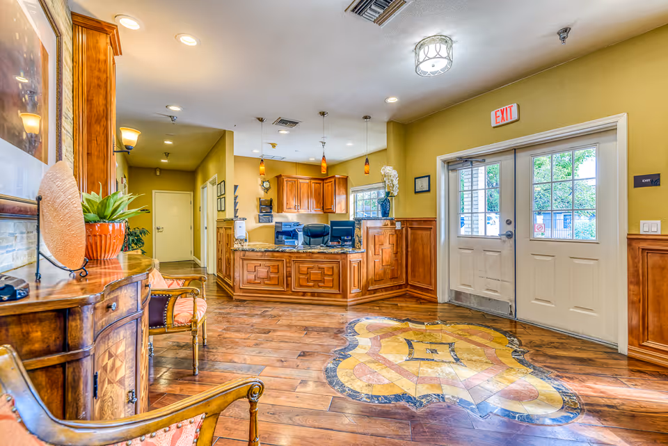 Reception area of the Cottages at Peoria facility featuring a wooden front desk with computer monitors, pendant lights hanging from the ceiling, wooden flooring with a decorative inlay, a double glass exit door, and seating with upholstered chairs and a wooden side table with a potted plant.