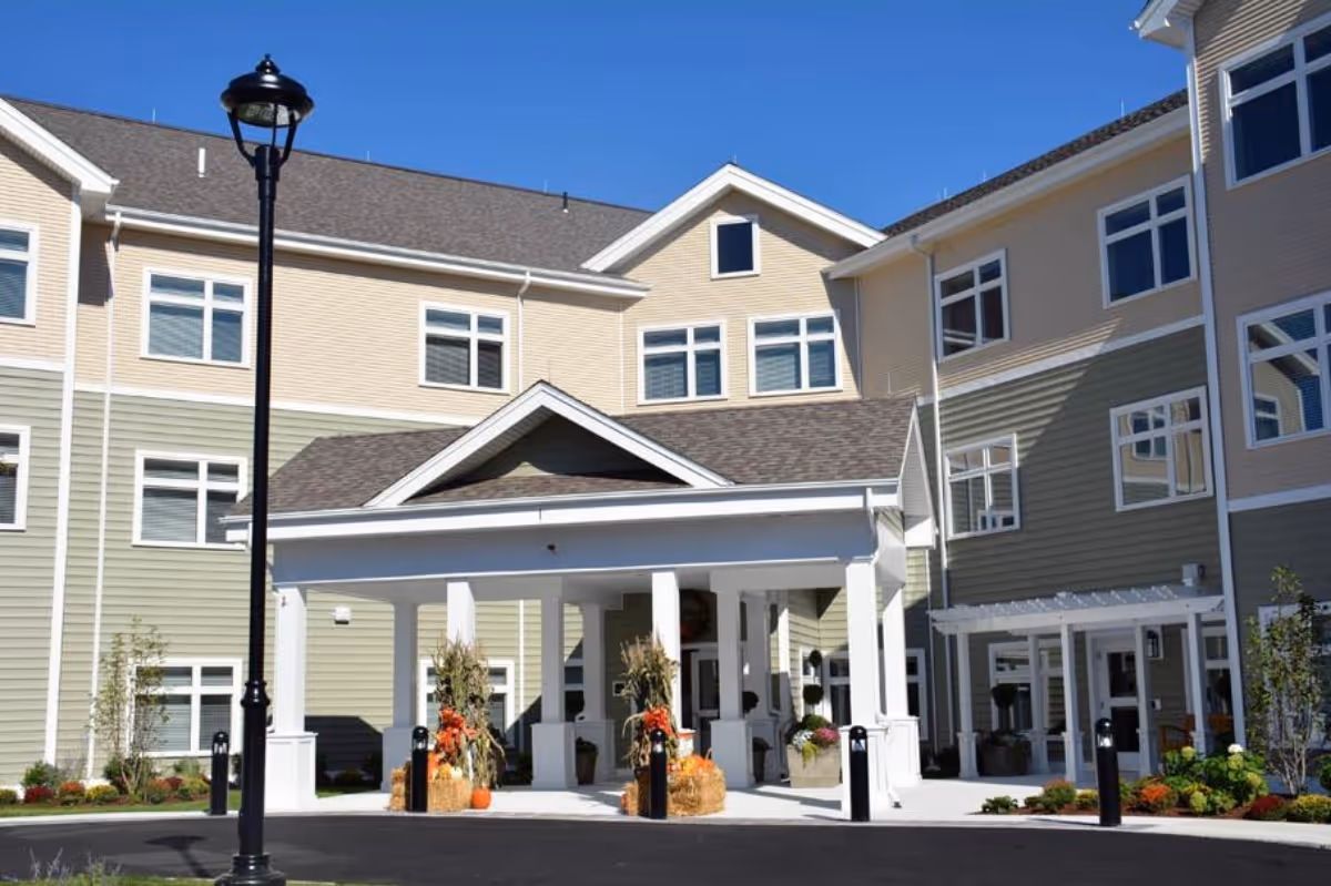 Exterior view of Benchmark Senior Living at Woburn showing a multi-story building with beige and green siding, white trim, and a covered entrance decorated with autumn-themed plants and pumpkins under a clear blue sky.