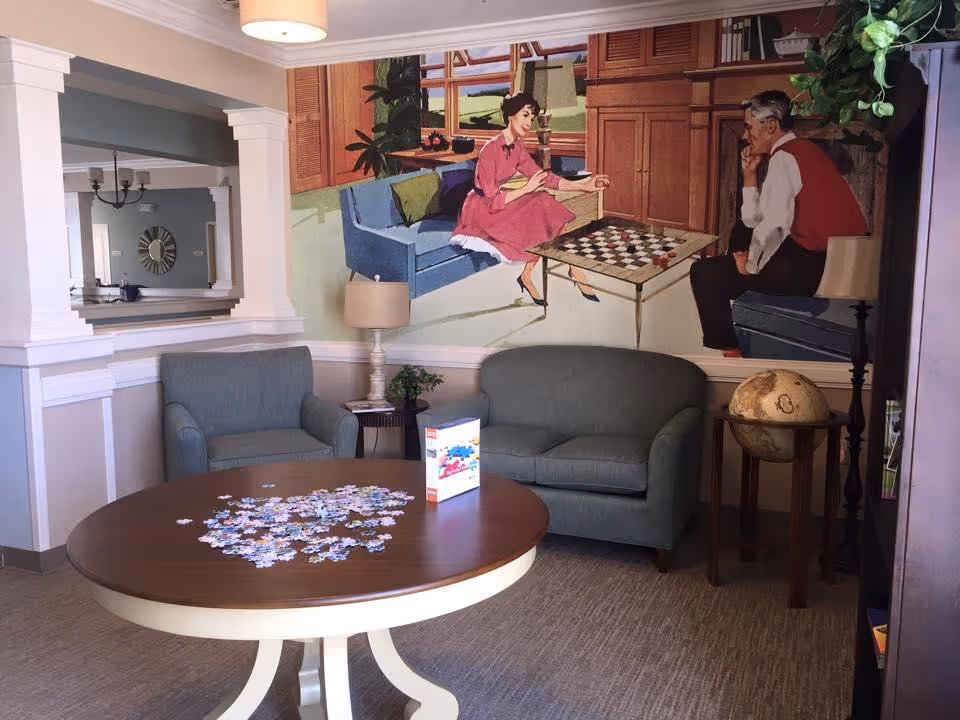 A cozy sitting area with a round wooden table in the center covered with scattered puzzle pieces and a puzzle box. Behind the table are a blue armchair and a blue loveseat. On the wall above the loveseat is a large mural depicting a man and woman playing checkers. There is a globe on a small wooden stand to the right and a lamp on a side table between the armchair and loveseat. The room has light-colored walls with white trim and a large mirror on the left wall.
