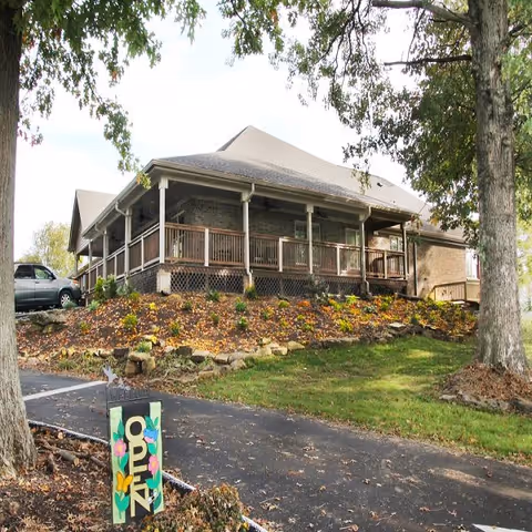 Single-story brick building with a wraparound porch on a landscaped rise, a driveway in front and a colorful 'OPEN' sign near the foreground.
