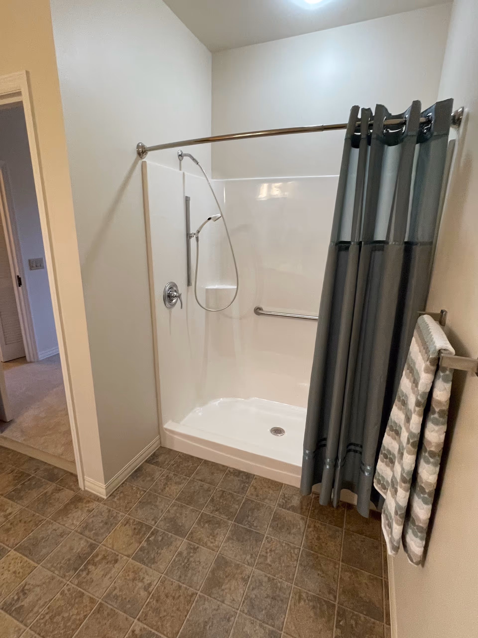 Accessible walk-in shower with a handheld showerhead, grab bar, gray curtain, and patterned towel in a bathroom with tiled floor.
