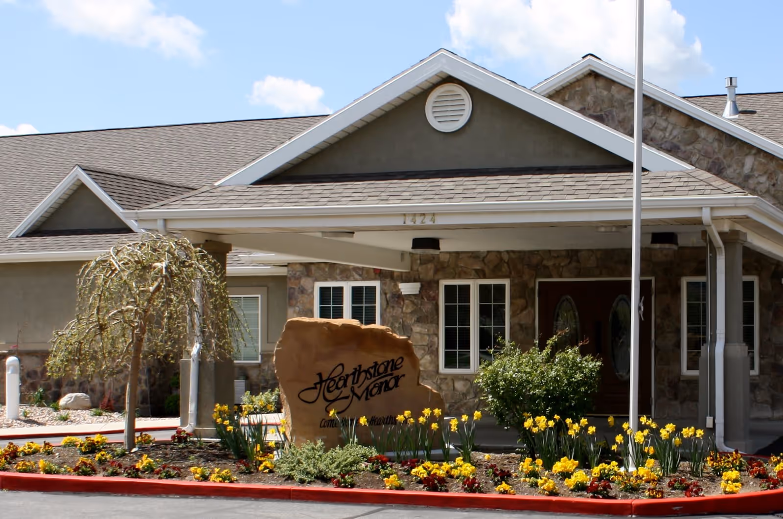 Exterior view of Hearthstone Manor, showing the entrance with a covered porch, stone facade, and a landscaped flower bed with yellow and red flowers in front of a large stone sign displaying the facility's name.