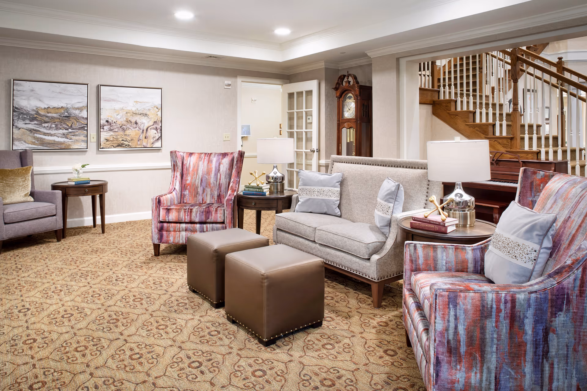 A cozy living room area with patterned carpet, two colorful armchairs with pillows, a gray sofa with pillows, two brown ottomans, side tables with lamps and books, abstract paintings on the wall, a grandfather clock, and a wooden staircase in the background.