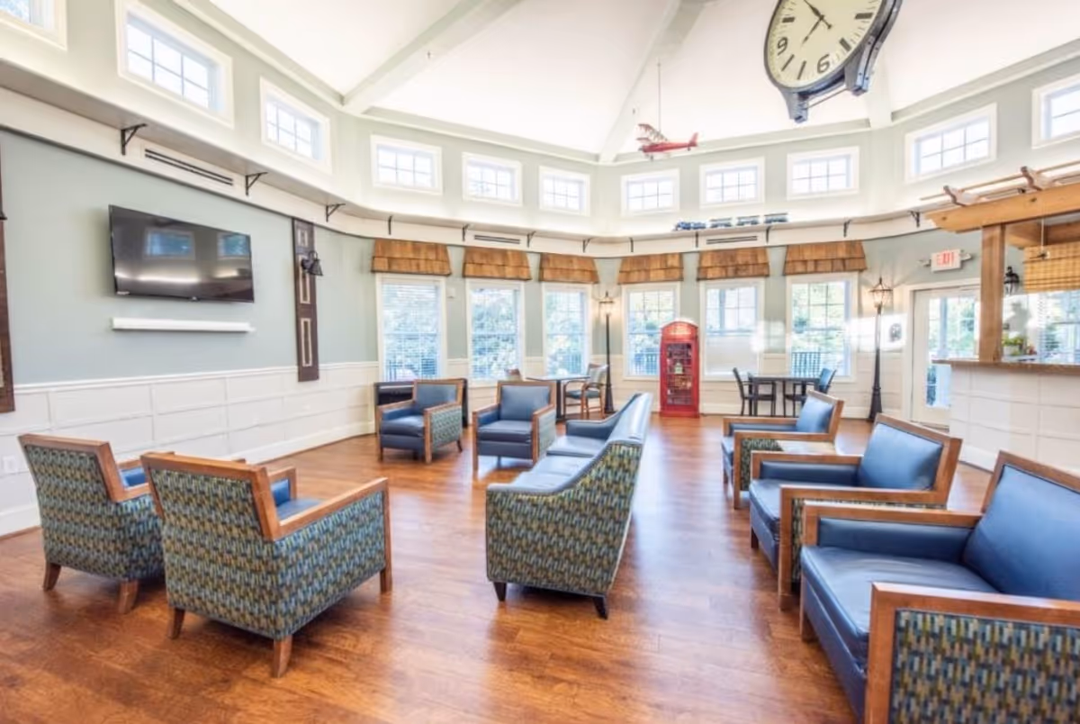 A spacious senior living facility common area with multiple blue cushioned armchairs and sofas arranged on a wooden floor. The room features large windows with brown blinds, a wall-mounted TV, a large clock hanging from the ceiling, and a red phone booth in the corner. The walls are painted light green with white wainscoting, and the ceiling has multiple small windows near the top allowing natural light to fill the room.