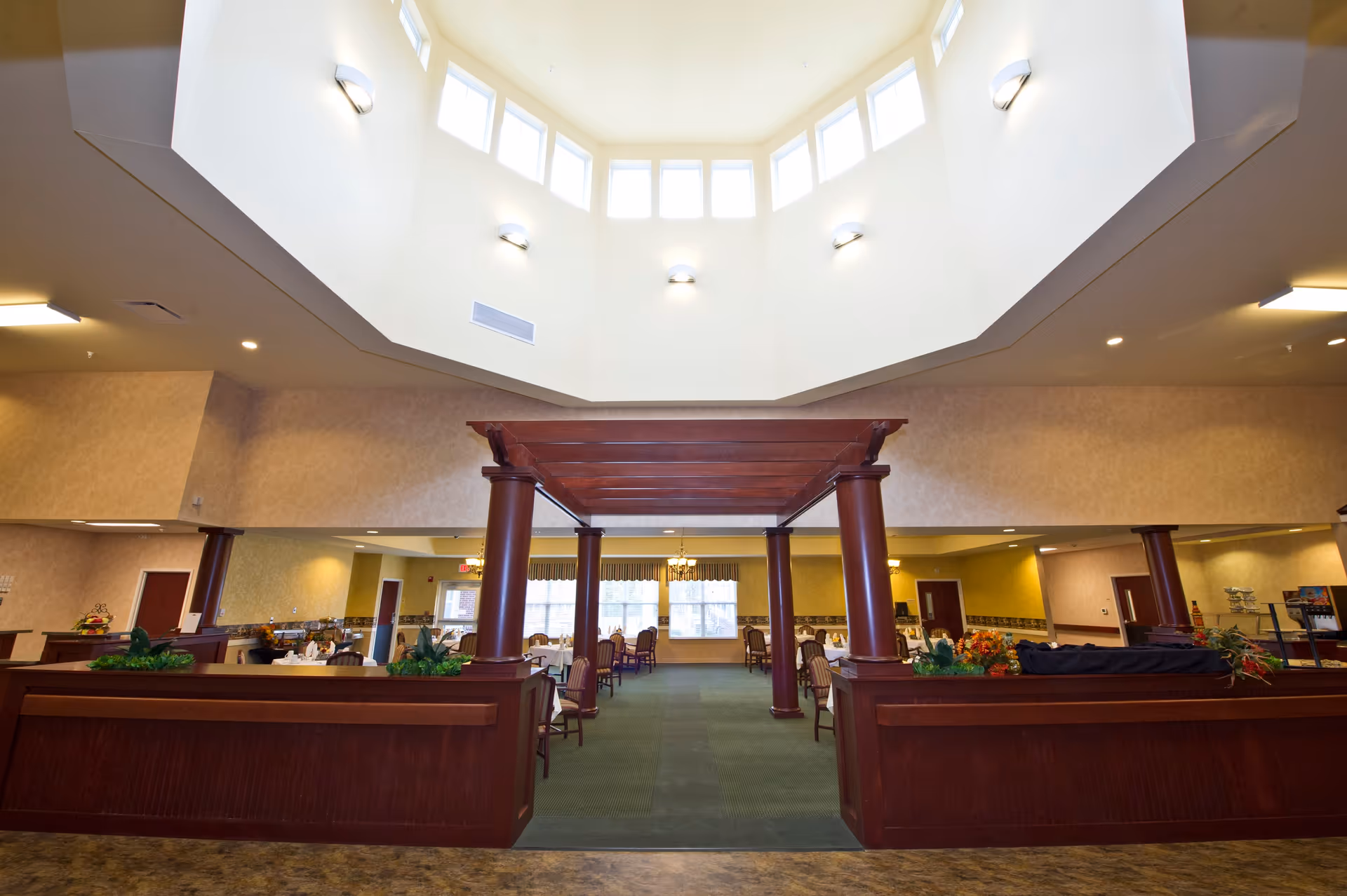 Interior view of a spacious dining room in a senior living facility with a high ceiling featuring multiple windows near the top, wooden columns and beams, and tables and chairs arranged for dining. The room is well-lit with wall sconces and chandeliers, and decorated with plants and floral arrangements.