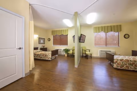Shared resident room with two beds, chairs, and windows in a senior facility.