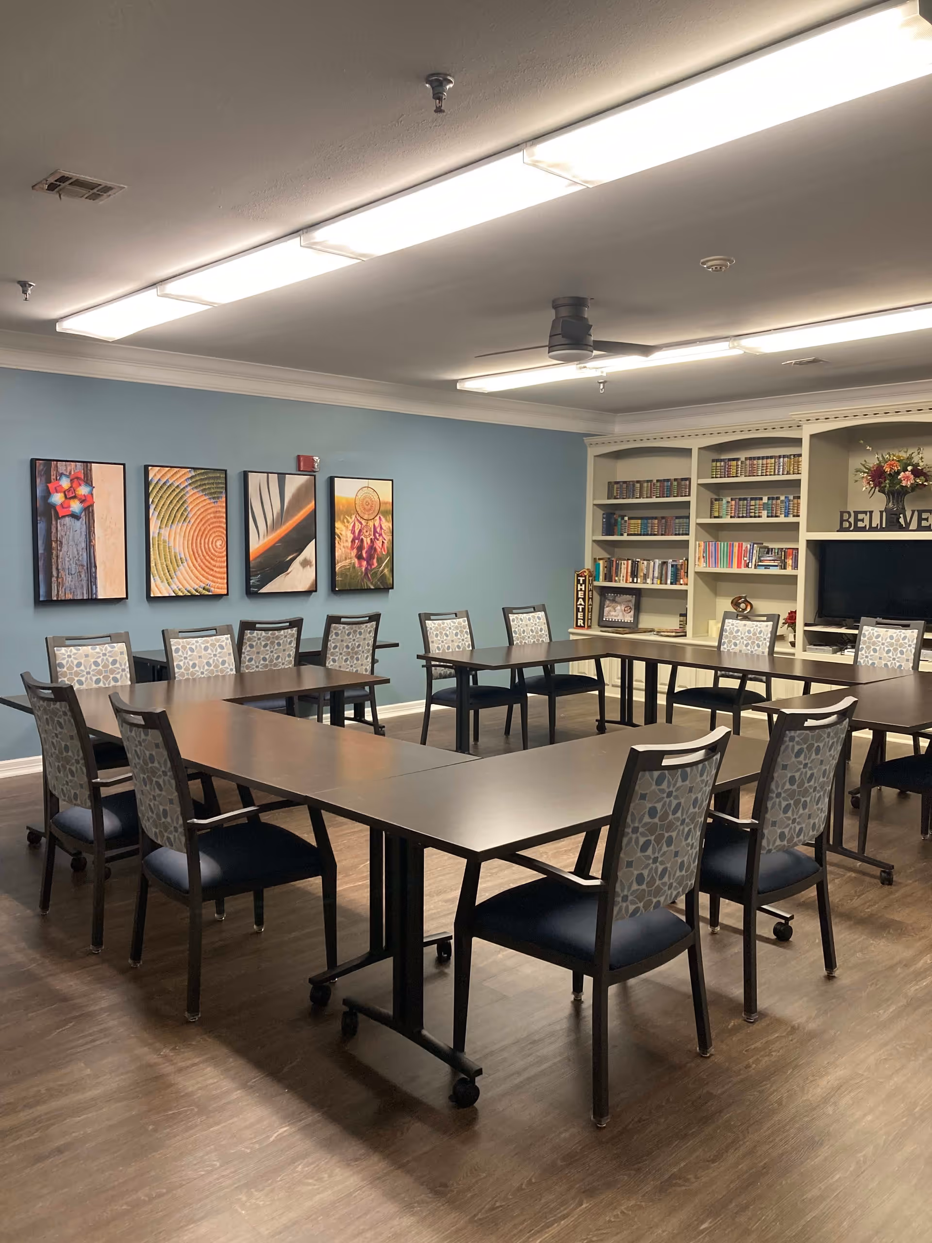 A well-lit room with tables arranged in a U-shape surrounded by chairs with patterned backs and dark seats. The walls are painted light blue and decorated with four colorful framed artworks. There is a large white bookshelf filled with books and decorative items, including a vase with flowers and a sign that says 'BELIEVE'. The floor is wooden, and the ceiling has fluorescent lights and a ceiling fan.