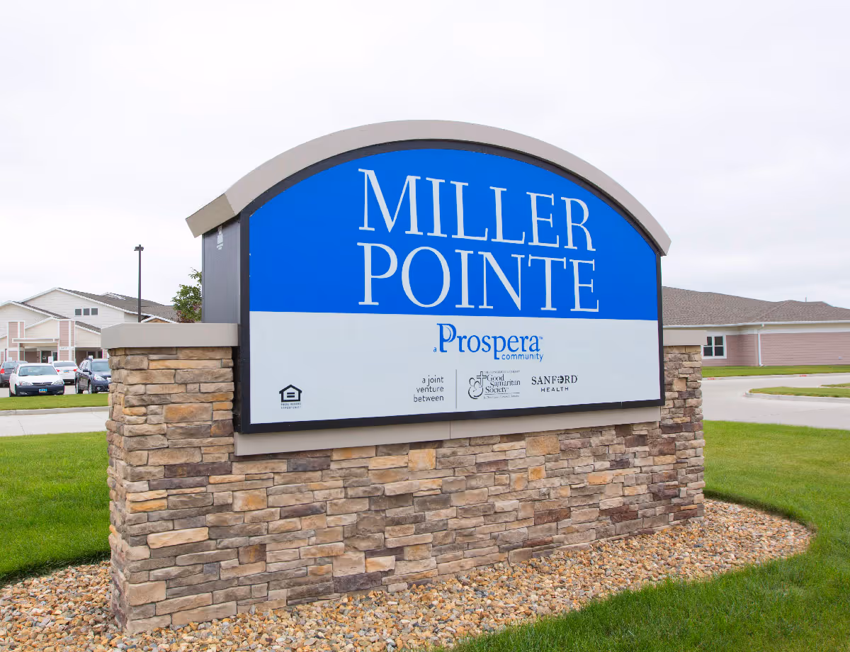 Outdoor stone and concrete sign for Miller Pointe, a Prospera community, a joint venture between Good Samaritan Society and Sanford Health, with residential buildings and parked cars in the background.
