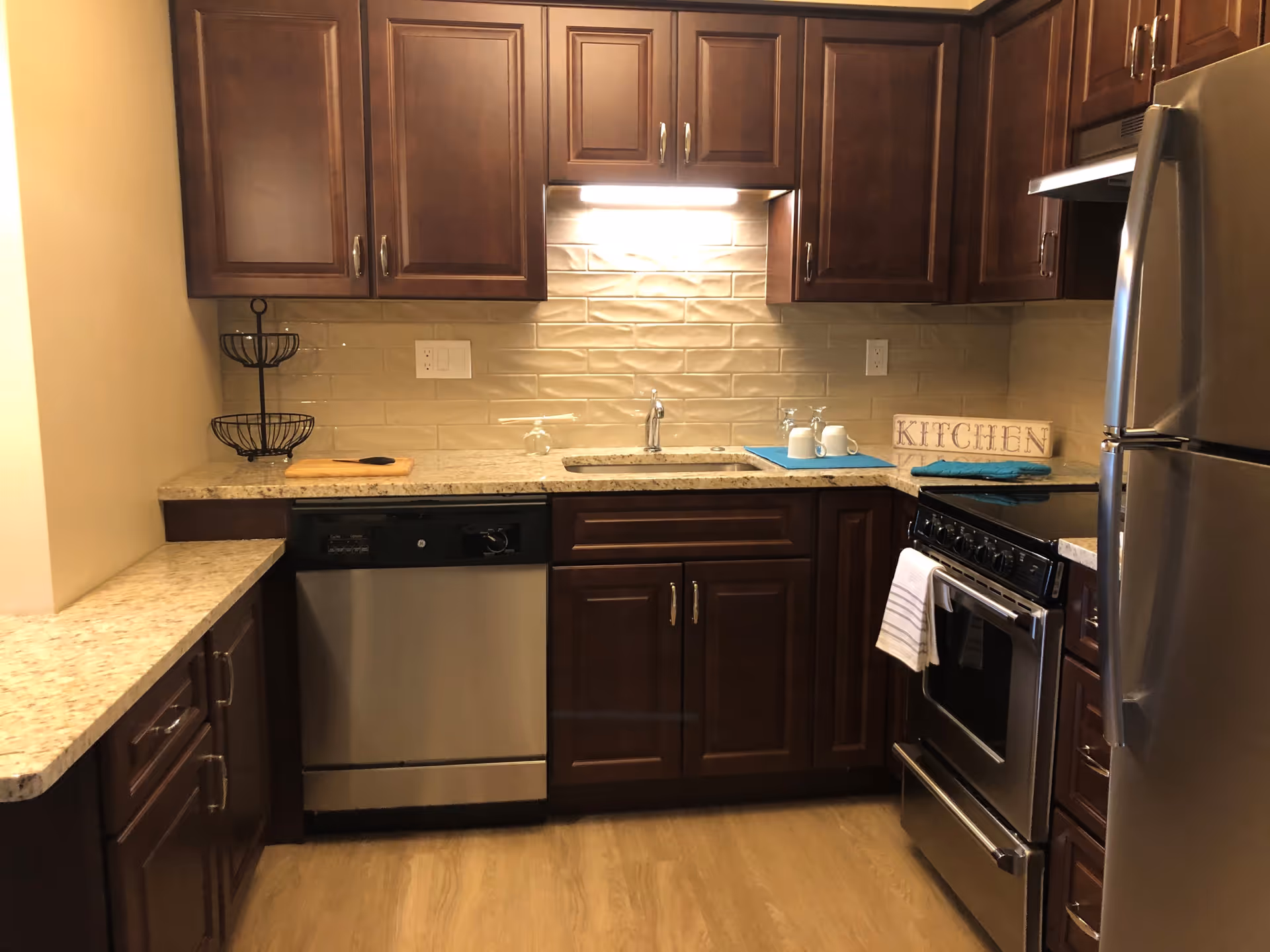 A kitchen with dark wooden cabinets, granite countertops, a stainless steel dishwasher, stove, and refrigerator. There is a sink under a cabinet light, a two-tiered fruit basket on the left countertop, and a decorative sign that says 'KITCHEN' on the right countertop next to two mugs and a blue mat.