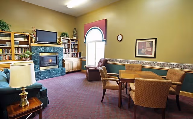 Common lounge with a fireplace, TV, bookshelves, armchairs and a table with chairs by a window.