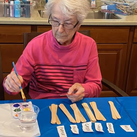 An elderly woman wearing glasses and a pink striped shirt is sitting at a table painting with a brush. In front of her are several wooden craft sticks and small pieces of paper labeled with colors such as purple, blue, green, yellow, orange, and red. There is a cup of water and a paint palette with various colors on the table. The background shows a kitchen counter with a sink and various items.