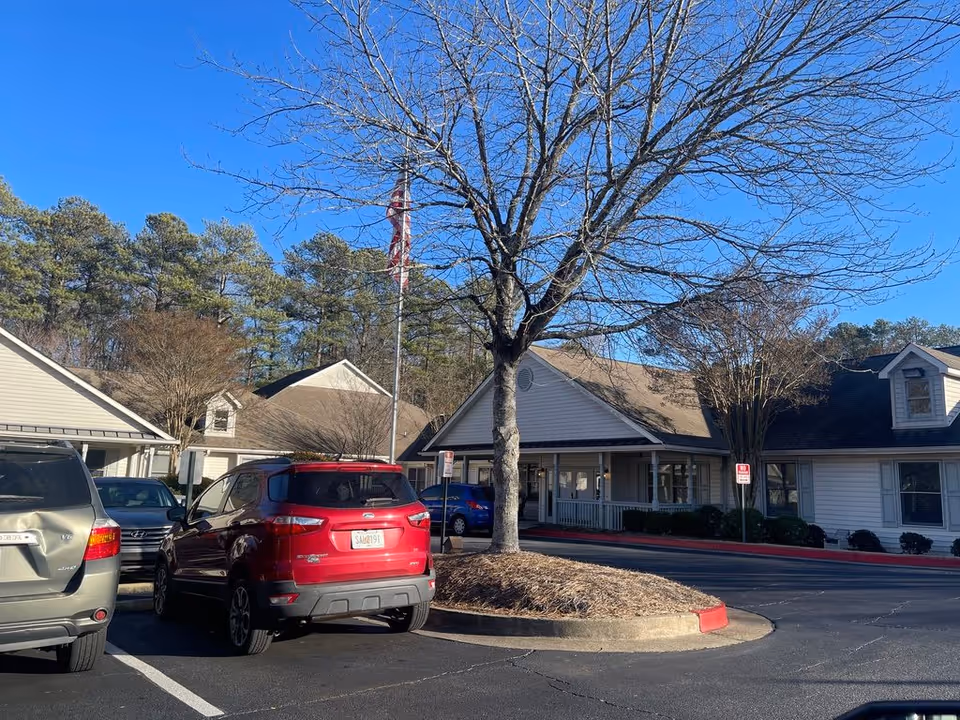 Parking lot with several parked cars, a leafless tree and flagpole in front of the single-story entrance of a senior living building under a clear blue sky.