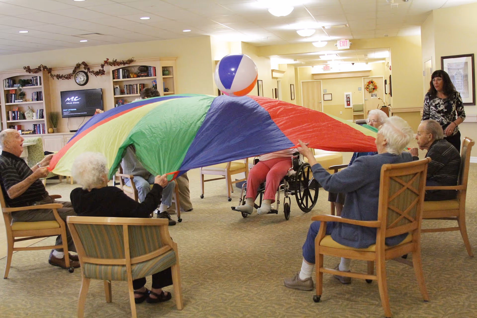 A group of elderly people sitting in chairs in a common area, playing with a colorful parachute and a beach ball. A woman stands nearby watching them. The room has bookshelves, a television, and warm lighting.