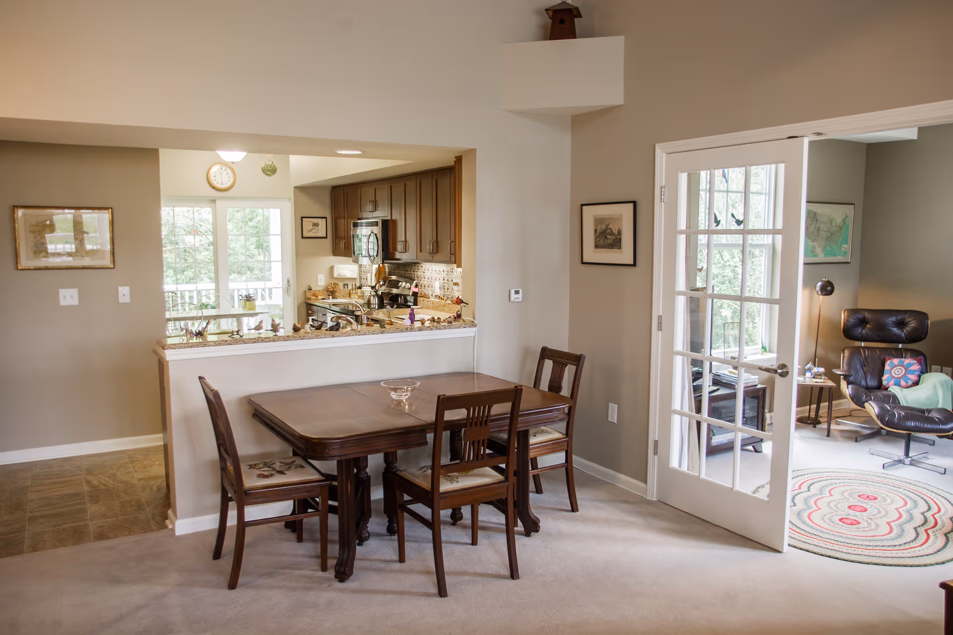 Interior view of a senior living facility showing a dining area with a wooden table and four chairs. The dining area is adjacent to a kitchen with wooden cabinets, a microwave, and a stove. To the right, there is a room with glass French doors, featuring a black leather lounge chair, a side table with a lamp, and a round rug on the floor. The walls are painted beige, and there are framed pictures hanging on them.