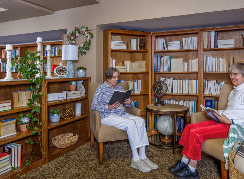 Two elderly women sitting in armchairs in a cozy library or reading room. One woman is wearing a blue patterned shirt and white pants, reading a book, while the other woman is wearing a white top and red pants, smiling and holding a book. The room has wooden bookshelves filled with books, decorative plants, a globe, and various decorative items including candles and a clock.