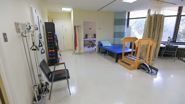 A rehabilitation room with exercise equipment including resistance bands, dumbbells, a therapy bed, parallel bars for walking exercises, and an exercise bike. There is a chair against the wall and a window with curtains letting in natural light.