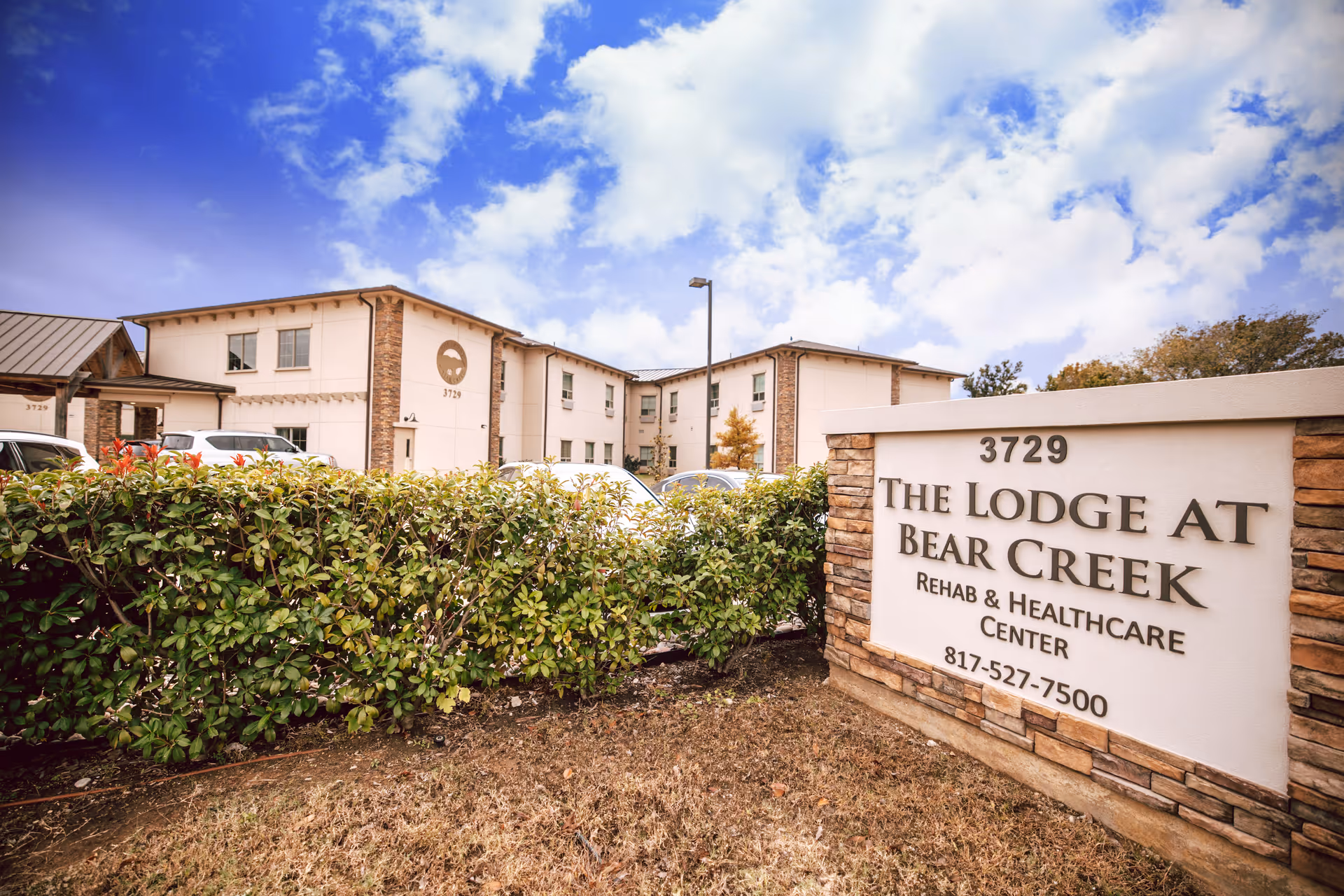 Exterior view of The Lodge at Bear Creek Rehab & Healthcare Center building with a sign displaying the name, address 3729, and phone number 817-527-7500. The building is two stories with beige walls and stone accents, surrounded by bushes and parked cars under a partly cloudy sky.