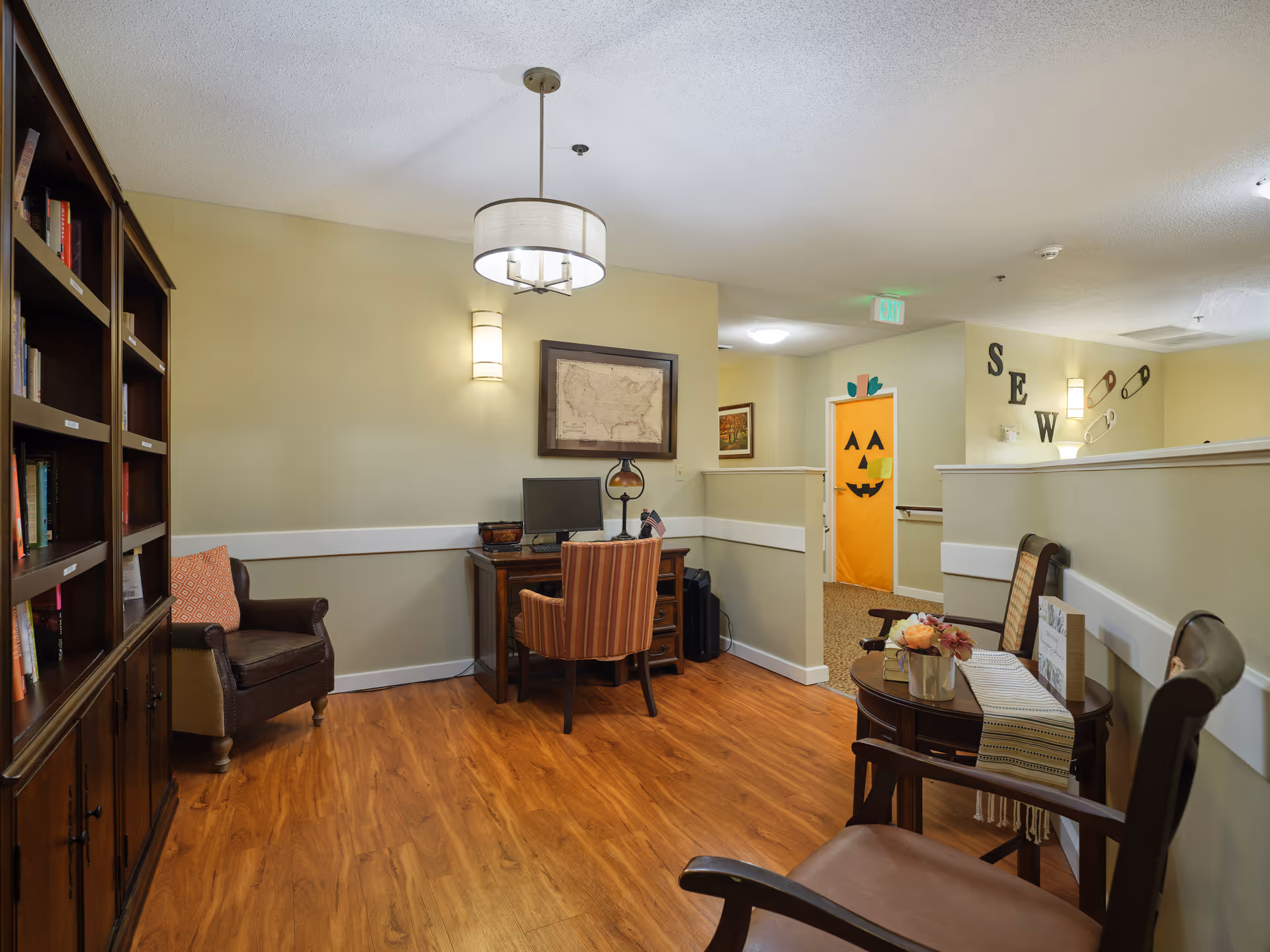 A cozy interior corner of a senior living facility with wooden flooring, a bookshelf filled with books, a desk with a computer and a striped chair, two wooden chairs around a small round table with a flower arrangement, and a hallway with a door decorated like a jack-o'-lantern pumpkin face. The wall has the word 'SEW' with large letters and oversized safety pins as decoration.