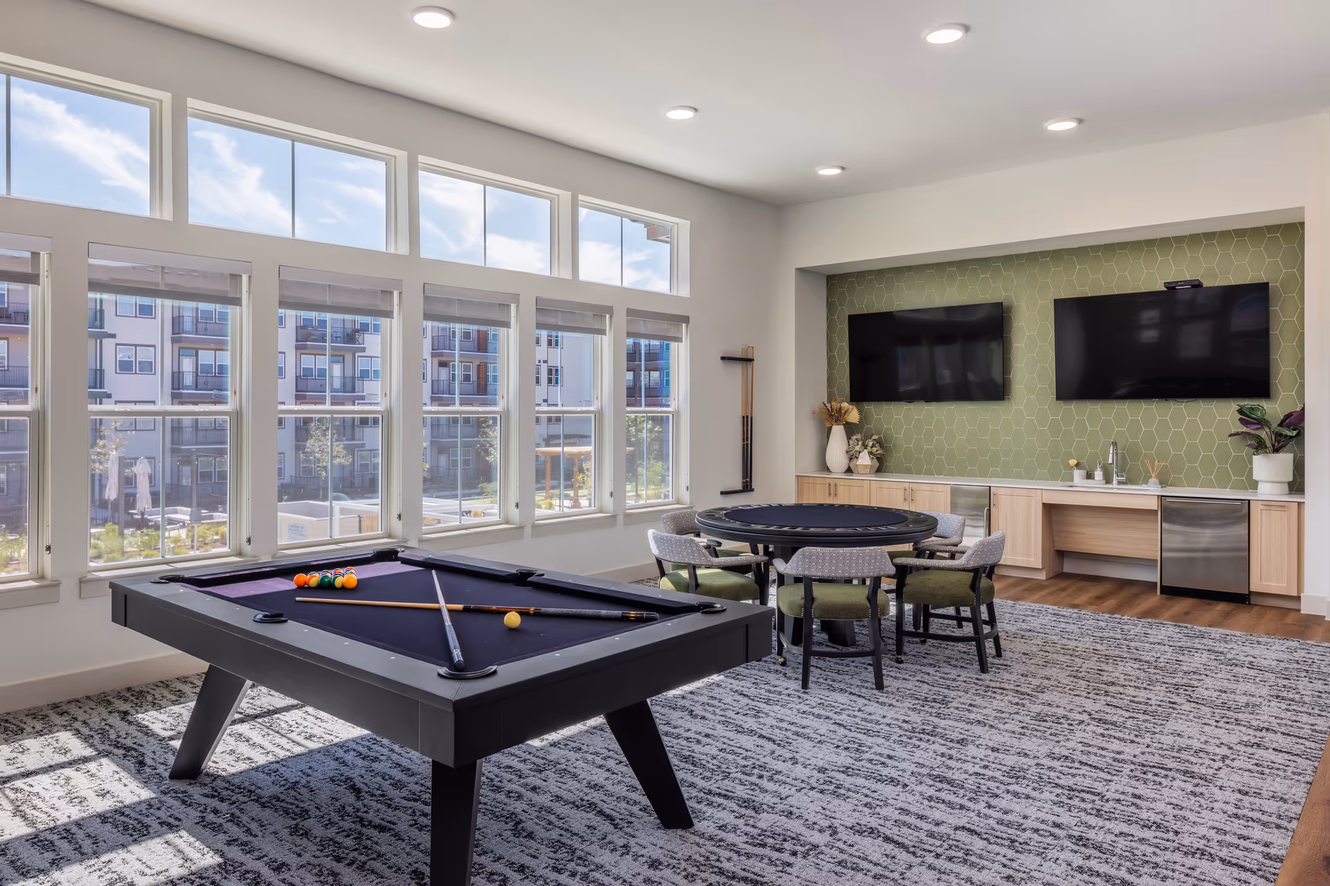 A bright game room featuring a billiards table with colorful balls, surrounded by large windows that let in natural light. There is a circular dining table with chairs, and two flat-screen TVs mounted on the wall.