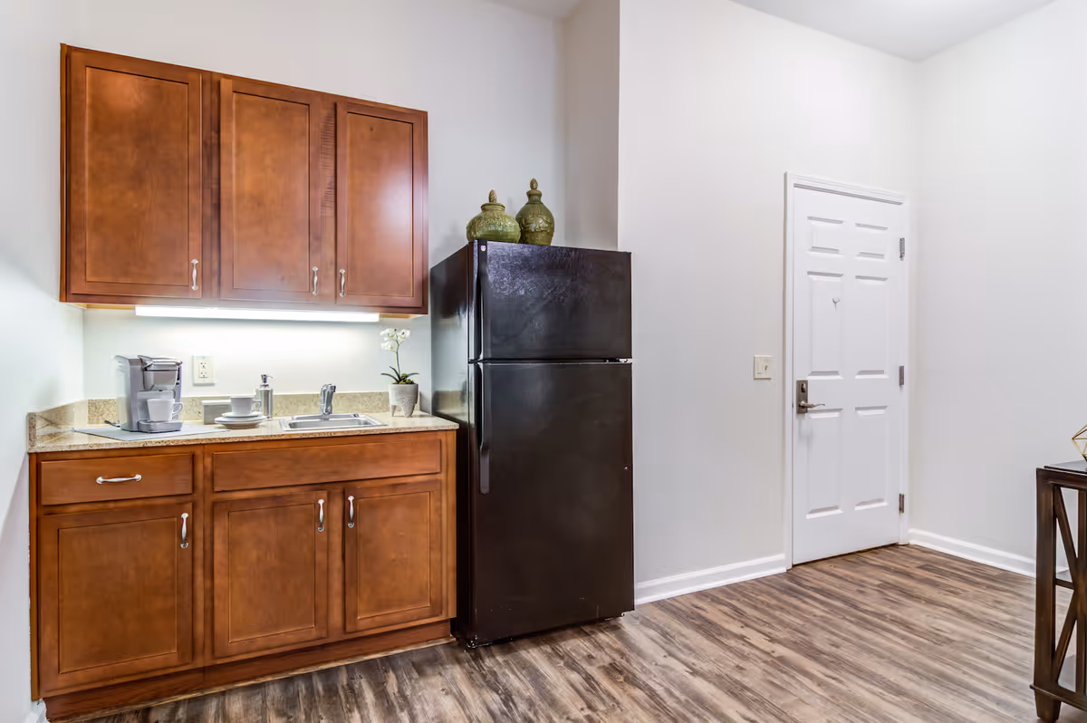 A small kitchenette area with wooden cabinets, a black refrigerator, a countertop with a coffee maker, cups, a small sink, and decorative green jars on top of the refrigerator. The floor has wood-style vinyl flooring and there is a white door to the right.