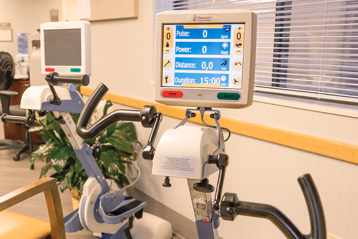 Two stationary exercise bikes with digital monitors in a well-lit room with a window and a plant in the background, likely part of a fitness or rehabilitation area.