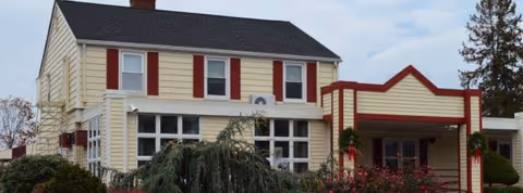 Two-story beige nursing home with red shutters and a covered entrance, surrounded by shrubs.