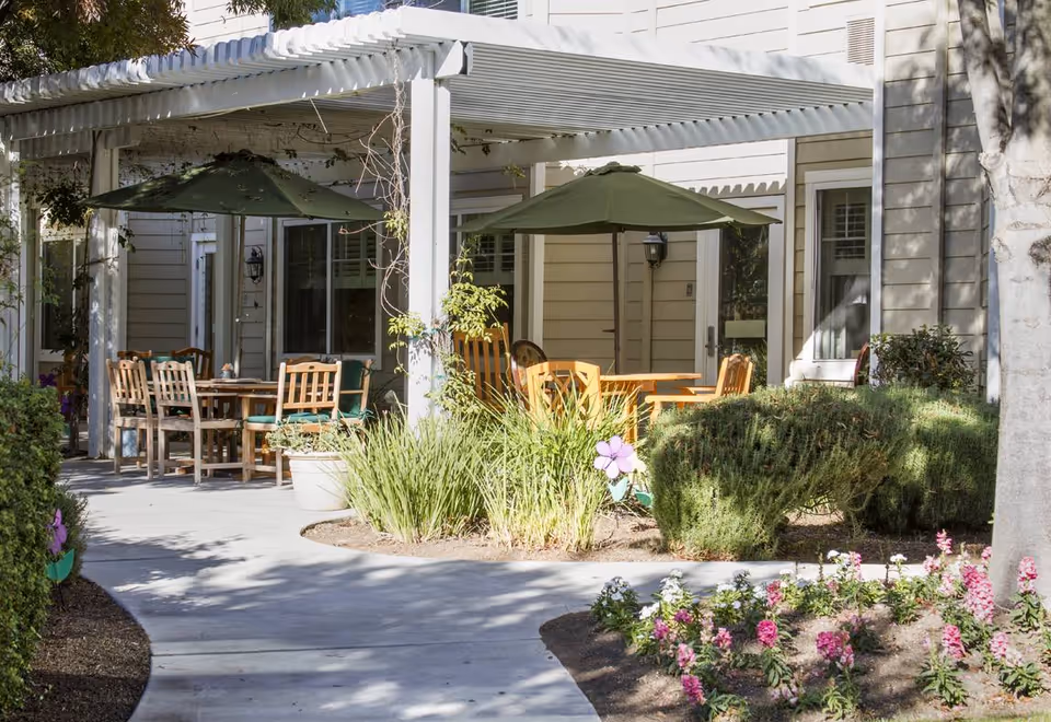 Outdoor patio area at The Grove Senior Living with wooden tables and chairs under green umbrellas, surrounded by plants, bushes, and flowers, adjacent to a beige building with windows and doors.