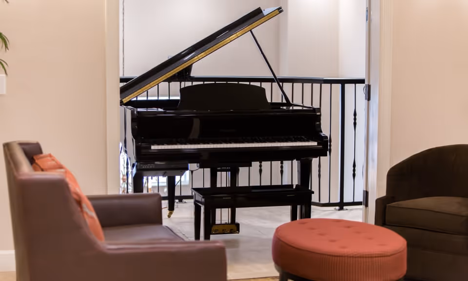 A cozy interior space featuring a black grand piano with its lid open, positioned near a railing. Surrounding the piano are comfortable seating options including a brown leather armchair with orange cushions, a dark upholstered chair, and a round red ottoman. The walls are light-colored, creating a warm and inviting atmosphere.