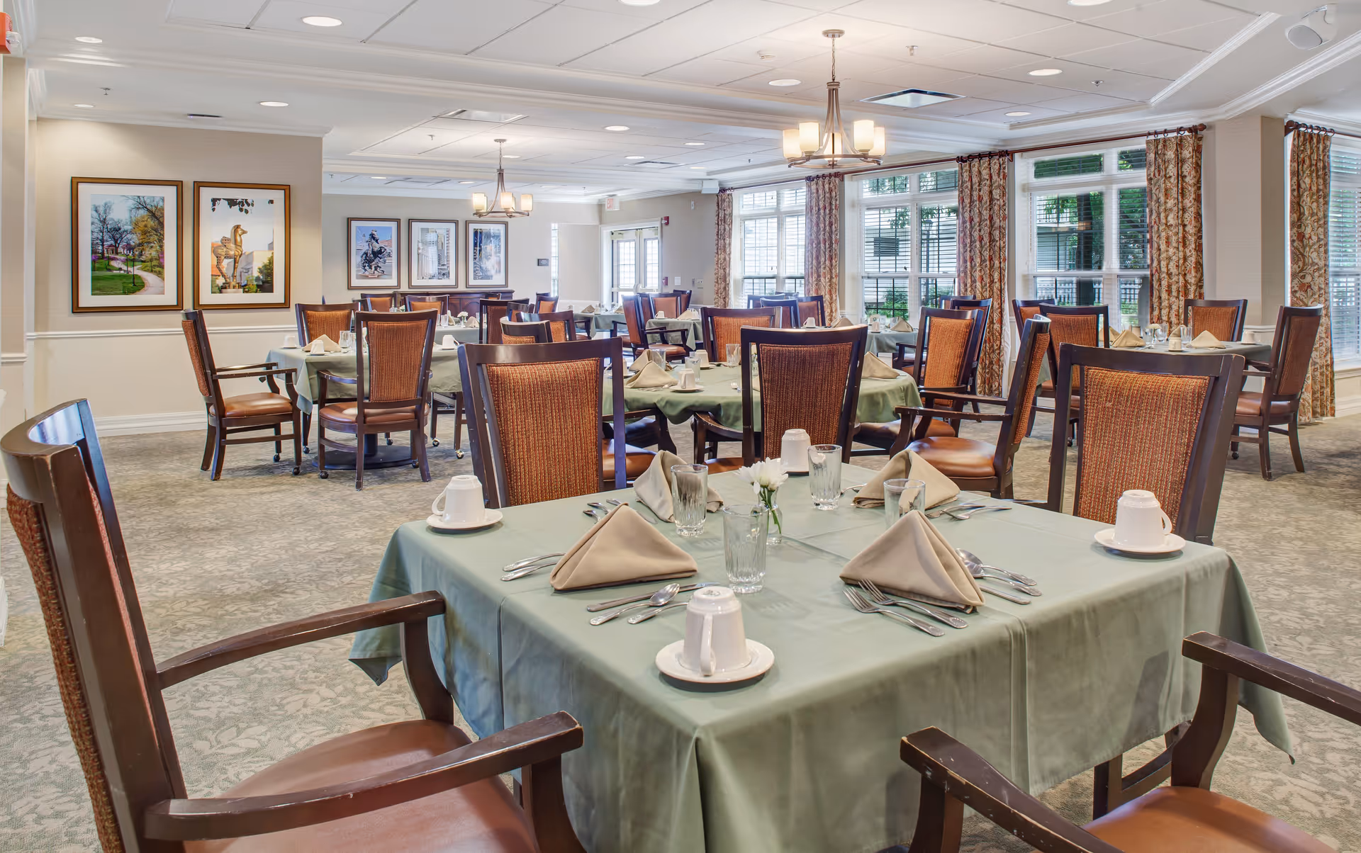 A spacious dining room with multiple tables covered in green tablecloths, each set with beige folded napkins, glasses, cups, and silverware. The room features large windows with patterned curtains allowing natural light to fill the space. The walls are decorated with framed artwork, and there are chandeliers hanging from the ceiling providing additional lighting.