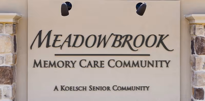 Sign on a beige wall with stone accents on the sides, displaying the text 'MEADOWBROOK MEMORY CARE COMMUNITY A KOELSCH SENIOR COMMUNITY'.