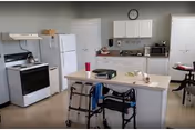 A bright kitchen area with white cabinets, a white refrigerator, a white stove, and a central island. On the island, there are a few items including a pink cup and some papers. A walker is positioned next to the island. In the background, there is a microwave on the countertop and a clock on the wall above the cabinets.