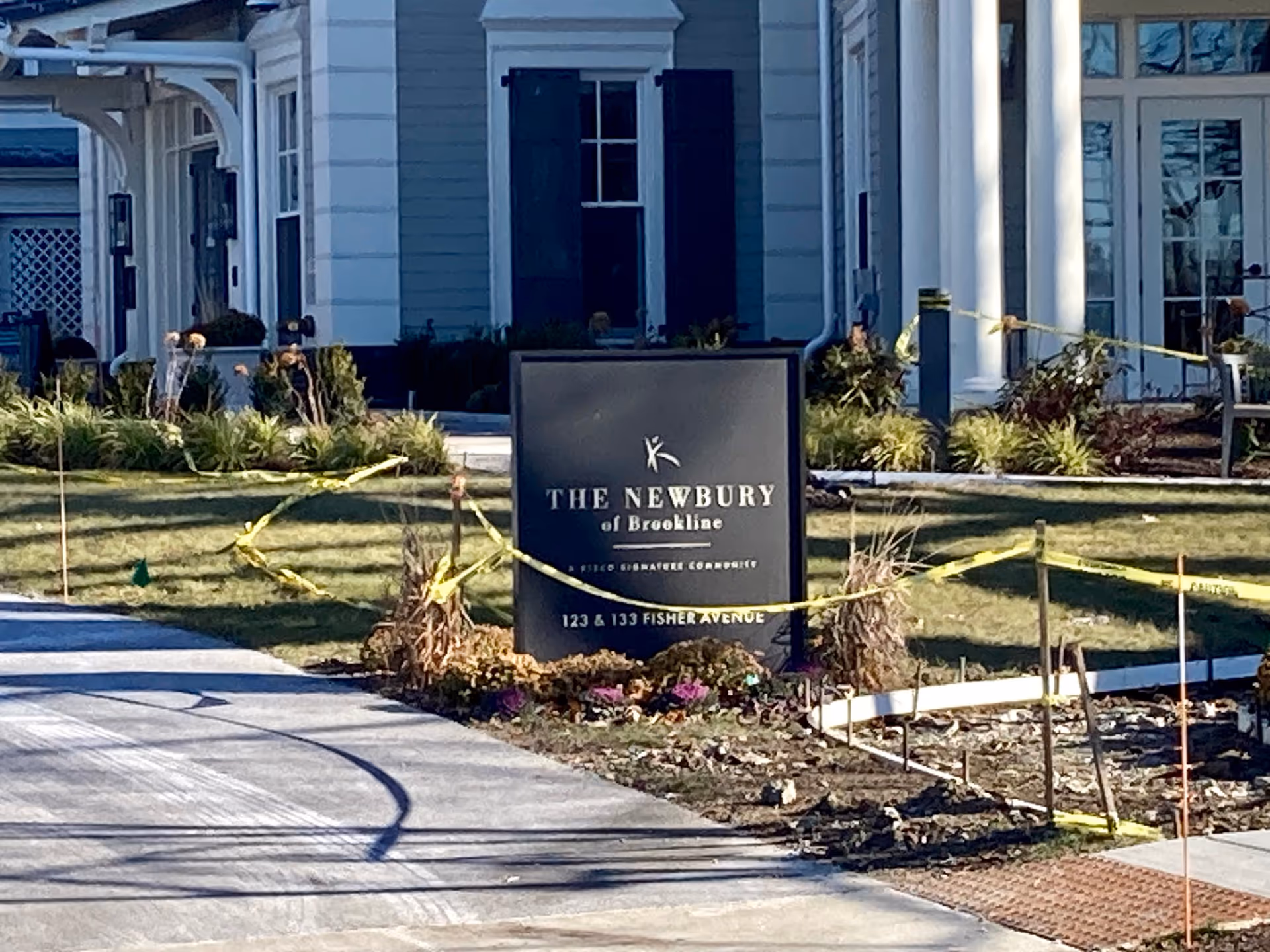 A black entrance sign reading The Newbury of Brookline stands on the lawn in front of a light-colored building, with yellow caution tape around the landscaping.
