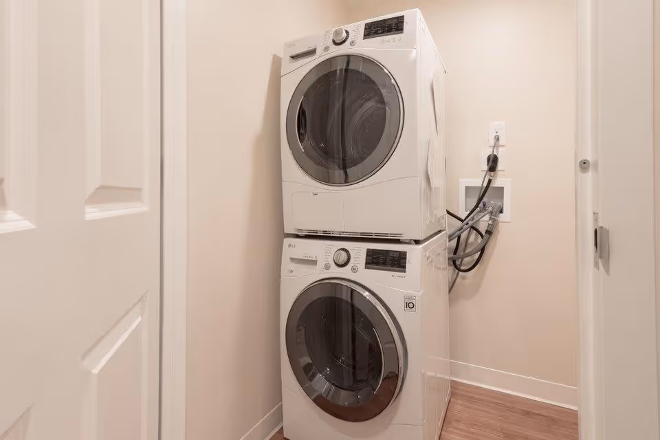 A small laundry area with a stacked white front-loading washer and dryer set against a beige wall. The machines are connected to water and power outlets on the right side. The floor is wood, and there is a white door partially visible on the left.