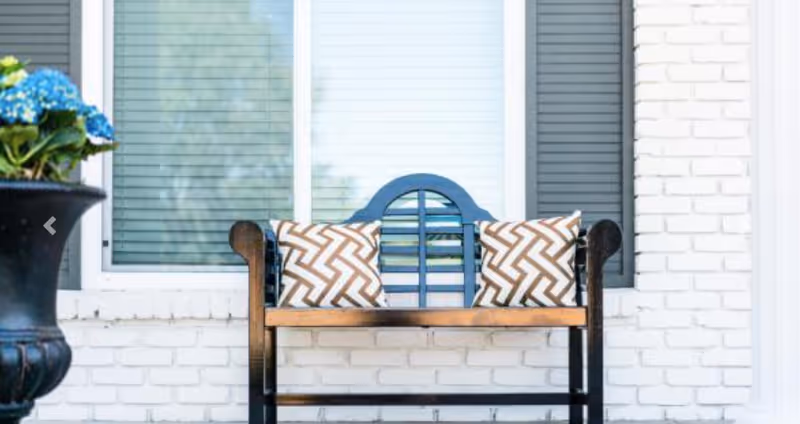Wooden bench with patterned pillows on a white brick porch in front of a window and a planter with blue flowers.