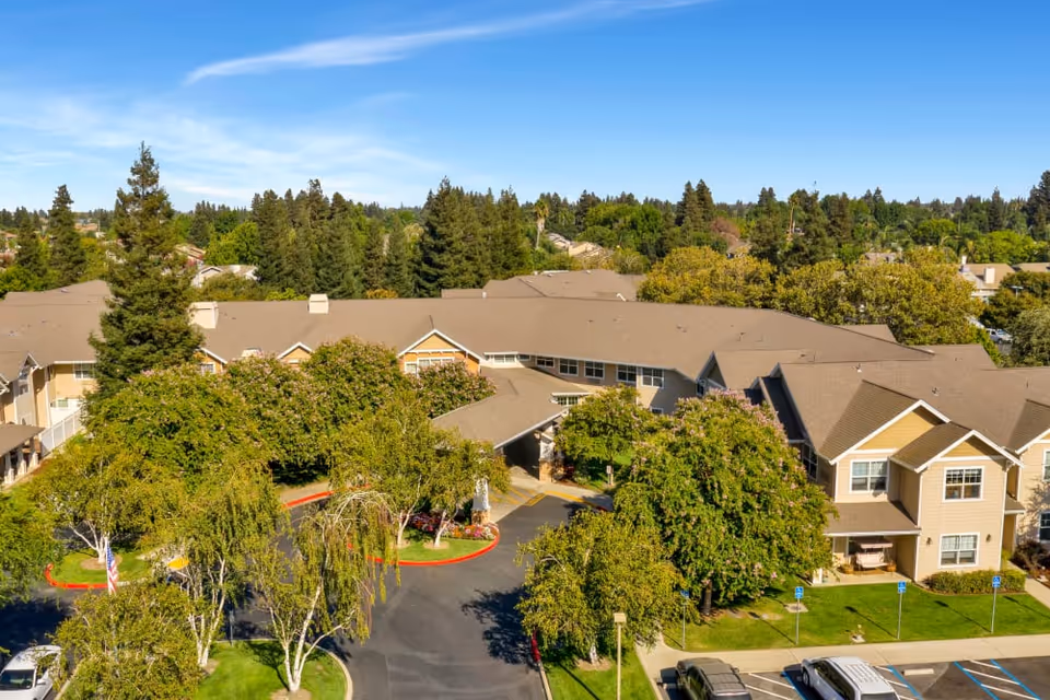 Aerial view of Dale Commons senior living facility surrounded by trees and greenery under a clear blue sky, with a parking lot visible in the foreground.